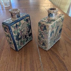 May include: Two rectangular ceramic bottles with floral and figure designs. The bottles have a beige background with blue, green, and purple floral patterns. The rims and necks of the bottles are a darker brown color.