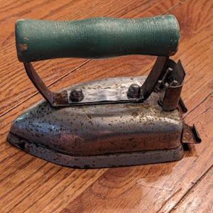 May include: Vintage metal iron with a green wooden handle. The iron has a worn, rusty finish and is labeled "The Perfect Iron".