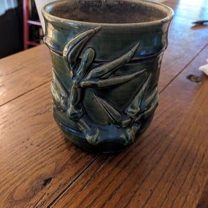 May include: A green ceramic planter with a raised design of bamboo leaves. The planter has a slightly textured surface and a brown rim.