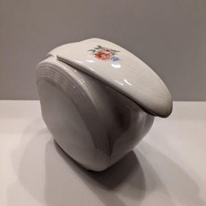 May include: White ceramic pitcher with a floral design on the lid. The pitcher has a rounded shape and a slightly textured surface.