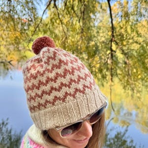 Op de afbeelding: Een gebreide wintermuts met een bruin en crèmekleurig zigzagpatroon en een bijpassende pompon. De muts wordt gedragen met een zonnebril en een kleurrijke trui. De achtergrond toont een wazig zicht op een meer en bomen.