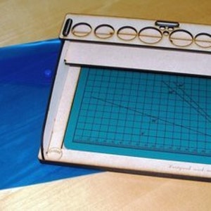 May include: A wooden paint organizer with a built-in cutting mat. The organizer has 10 slots for paint bottles and a sliding lid. The cutting mat is green and has a grid pattern. The organizer is labeled "Designed and made in England."