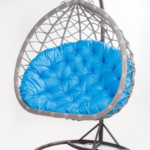 May include: A gray wicker egg chair with a bright blue cushion. The chair hangs from a curved metal stand. The cushion is tufted and appears soft. The chair is designed for relaxation and outdoor use.