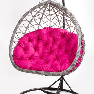 May include: A gray wicker hanging chair with a bright pink cushion. The chair is suspended from a black metal frame and has a circular base. The cushion is tufted and adds a pop of color to the neutral tones of the chair.