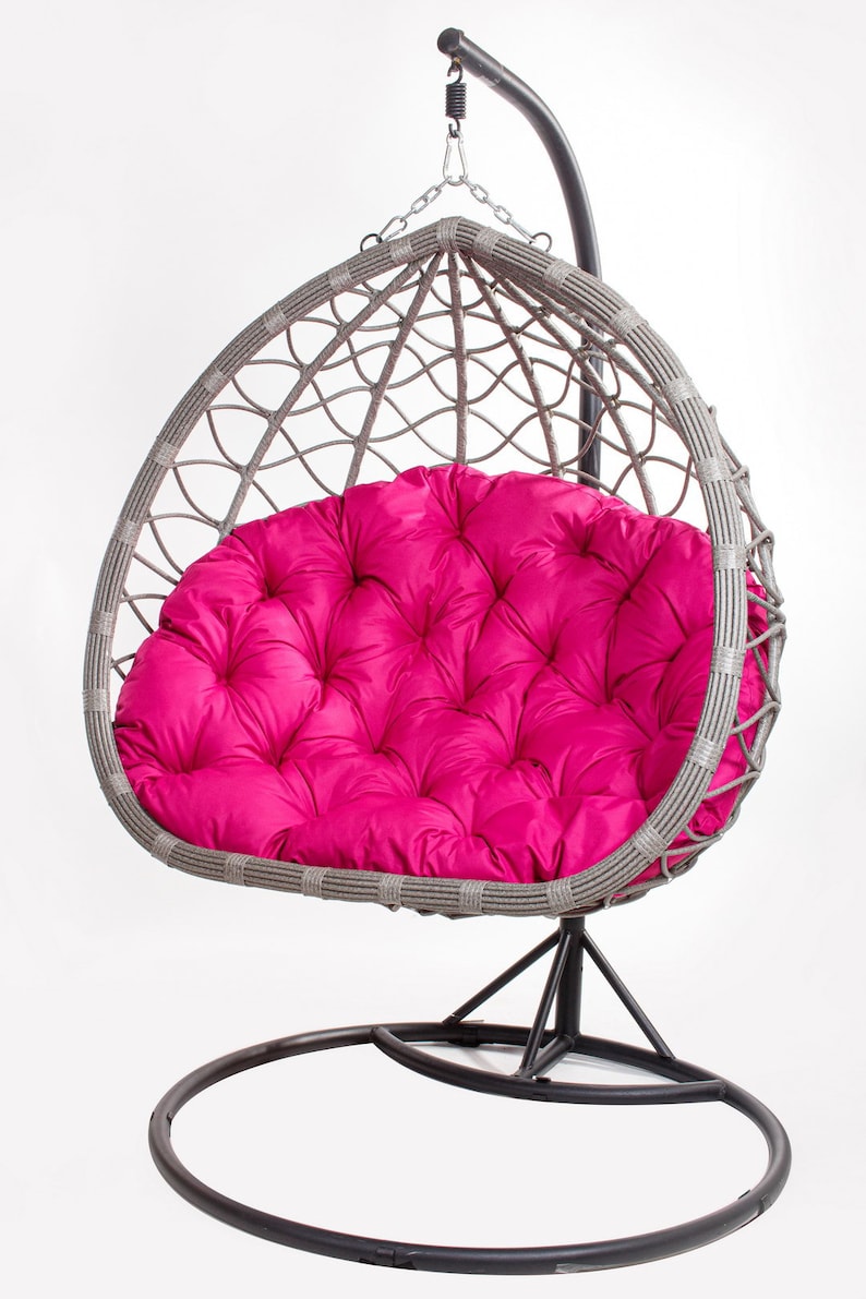 May include: A gray wicker hanging chair with a bright pink cushion. The chair is suspended from a black metal frame and has a circular base. The cushion is tufted and adds a pop of color to the neutral tones of the chair.