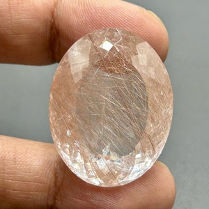 Può includere: Una gemma trasparente di forma ovale con taglio sfaccettato. La pietra ha un colore marrone chiaro e contiene molte inclusionesioni sottili di colore marrone rossiccio.