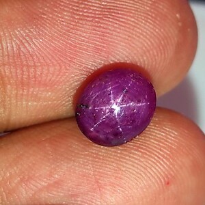 May include: A close-up of a polished, oval-shaped, deep magenta gemstone. The gem features a six-rayed star pattern across its surface. The stone is held between two fingers, highlighting its color and shape.