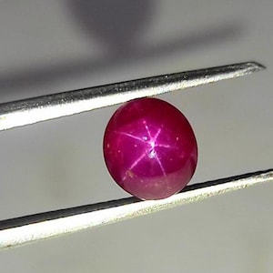 May include: A close-up of a red star ruby gemstone held in tweezers. The ruby is round and has a star-shaped pattern in the center.