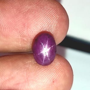 Puede incluir: Una piedra preciosa de rubí estrella de color púrpura oscuro con una estrella de seis puntas en el centro. El rubí tiene forma ovalada y una superficie lisa y pulida.