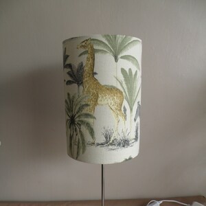 May include: A chrome table lamp with a white fabric lampshade featuring a giraffe and palm trees. The lampshade has a green, brown, and yellow colour scheme.