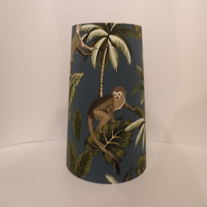 May include: A teal lampshade with a tropical print of monkeys, palm trees, and large green leaves. The lampshade has a cylindrical shape, tapering slightly towards the top. The design features brown and tan monkeys against a teal background.