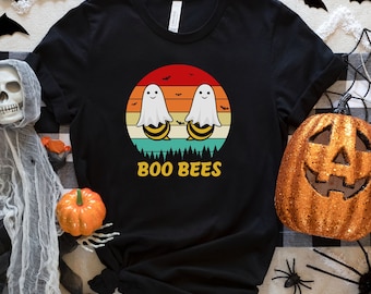 May include: Black t-shirt featuring a Halloween-themed graphic. The design includes two cartoon bees dressed as ghosts, set against a sunset backdrop with the text "BOO BEES". The shirt is styled with rolled-up sleeves.