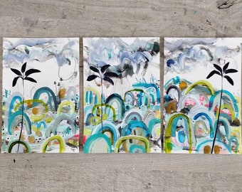 Op de afbeelding: Drie abstracte schilderijen met een landschapsthema. Elk schilderij heeft een blauw, groen en geel kleurenschema met een witte achtergrond. De schilderijen zijn wit ingelijst en worden getoond op een houten oppervlak.