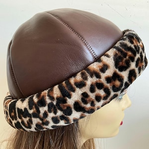 May include: A brown leather hat with a leopard print trim. The hat features a rounded crown and a soft, plush leopard print band around the brim. This hat is designed for cold weather wear.