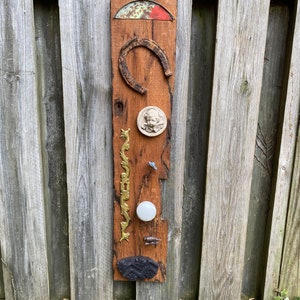 May include: A wooden board with various objects attached to it, including a horseshoe, a metal plate with a design, a small white knob, a gold-colored metal piece, and a black piece of wood.