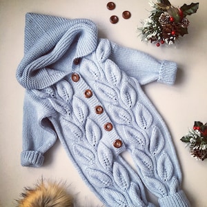 May include: A light blue knitted baby jumpsuit with a hood and leaf pattern. The jumpsuit has wooden buttons and is made of a soft, cozy material.