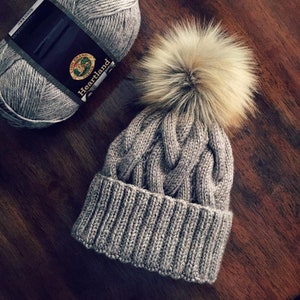 May include: A gray knitted hat with a cable knit pattern and a fluffy faux fur pom pom. The hat is made with Lion Brand Heartland yarn.