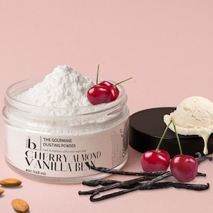 May include: A glass jar of white dusting powder, labelled "Cherry Almond Vanilla Bean", with fresh cherries, vanilla pods, and a scoop of ice cream. The jar contains 118 ml of powder. The background is pink.