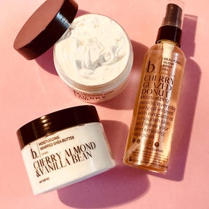 May include: A selection of beauty products on a pink surface. Features a jar of whipped shea butter with "Cherry Almond & Vanilla Bean" text, a jar of "Cherry Moisturizing Butter", and a bottle of "Cherry Glazed Donut" body oil.