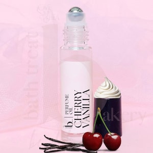 May include: A clear glass roller bottle with a silver roller ball. The bottle is labeled "Cherry Vanilla" and "Perfume Oil". The bottle is sitting on a pink surface with a small white cupcake and two red cherries.
