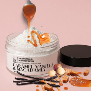 May include: A jar of "The Gourmand Dusting Powder" with caramel, vanilla, and macadamia flavour. Caramel sauce drips from a spoon onto the white powder. Vanilla beans, macadamia nuts, and caramel sweets are scattered around the jar.