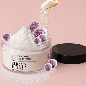 May include: A clear jar of white dusting powder labeled The Gourmand Dusting Powder Sugar Plum 4oz (118 ml) with a black lid. A gold spoon pours white powder into the jar, which is surrounded by several small, round, purple sugar-coated plums.