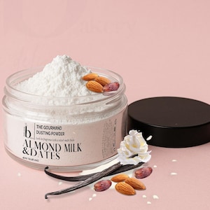 May include: A clear glass jar filled with white dusting powder, labeled "ALMOND MILK & DATES." The jar is open, with almonds and dates on top. A black lid sits beside the jar. Vanilla beans and whipped cream are arranged near the jar.