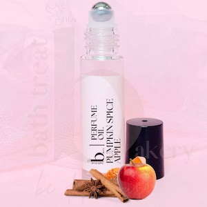 May include: A clear glass roller bottle with a black label that reads "PERFUME OIL by bath treat PUMPKIN SPICE APPLE". The bottle is sitting on a pink surface with cinnamon sticks, star anise, and a red apple.