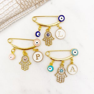 May include: Three gold safety pins with charms. Each pin has a hamsa hand charm, a blue evil eye charm, and a white enamel circle charm with a letter. The letters are L, P, and A.