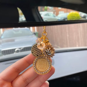 May include: Gold car charm with a circular pendant featuring Arabic calligraphy, surrounded by clear rhinestones. The charm also includes a smaller circular pendant with Arabic calligraphy, a clear rhinestone charm, and a charm with the Islamic symbol of faith.