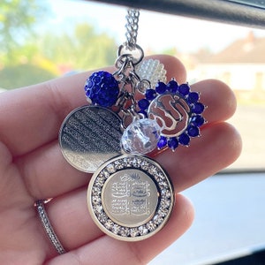 May include: A silver charm necklace with a variety of charms, including a round charm with Arabic script, a round charm with a blue crystal, a round charm with a clear crystal, and a charm with a blue crystal flower. The charms are attached to a silver chain.