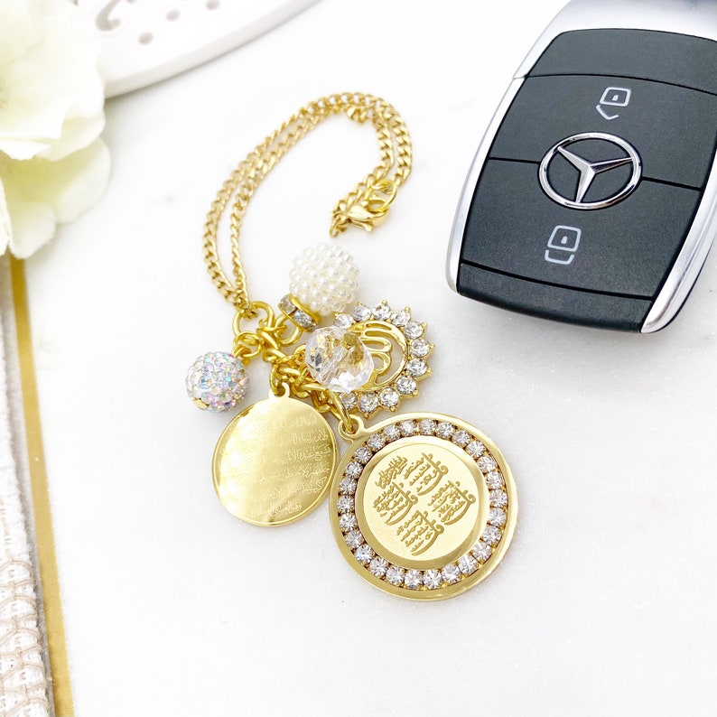 May include: Gold car charm with a round pendant featuring Arabic calligraphy, surrounded by clear rhinestones. The charm also includes a smaller round pendant with Arabic calligraphy, a crystal, a pearl bead, and a sparkly bead.