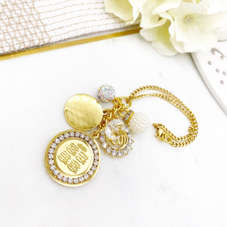 May include: Gold necklace with multiple charms, including a round charm with Arabic script, a round disc, a crystal charm, and a pearl charm.