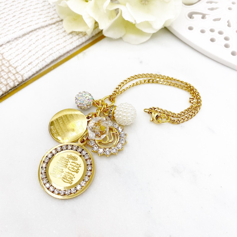 May include: Gold necklace featuring a selection of charms, including a round charm with Arabic script, a small flower charm, a pearl charm, and a sparkly charm. The necklace is displayed on a white surface.