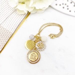 May include: Gold necklace with a charm featuring Arabic calligraphy surrounded by clear rhinestones. The necklace also has a round charm with a textured surface, a clear crystal charm, and two pearl charms.