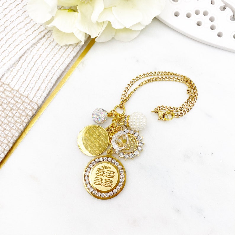 May include: Gold necklace with a charm featuring Arabic calligraphy surrounded by clear rhinestones. The necklace also has a round charm with a textured surface, a clear crystal charm, and two pearl charms.