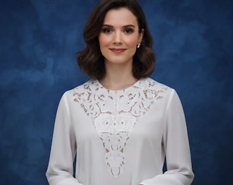 May include: A long-sleeved white blouse featuring a prominent V-shaped lace panel across the chest and shoulders. The lace displays a detailed floral pattern, while the rest of the garment is made of a smooth, solid white fabric with a round neckline.