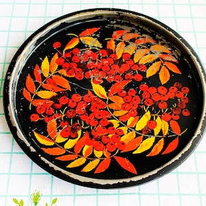 Puede incluir: Bandeja redonda de metal negro con un diseño pintado a mano de bayas rojas y hojas naranjas y amarillas. La bandeja tiene un aspecto ligeramente desgastado, con algo de desgaste de la pintura en los bordes. El diseño está centrado.