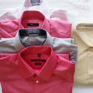 May include: A collection of folded button-down shirts in various colors, including pink, red, gray, and a light yellow. Some shirts have subtle patterns, and one is still in its original packaging. The shirts are neatly arranged, showcasing their collars and textures.
