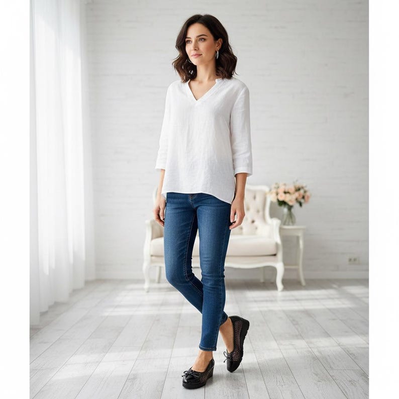 May include: A woman wearing a white V-neck blouse with three-quarter sleeves and dark blue jeans. She is also wearing black shoes. The setting is a bright room with a white brick wall and a white chair.