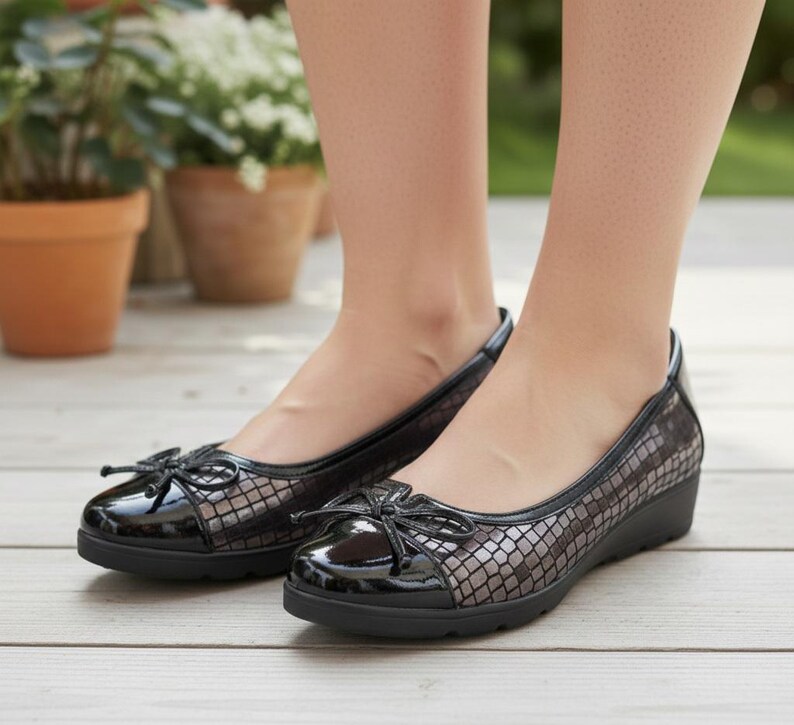 May include: Black ballet flats with a bow detail. The shoes have a glossy black toe cap and a textured, patterned design on the upper. The soles are black and slightly elevated. The shoes are designed for comfort and style.
