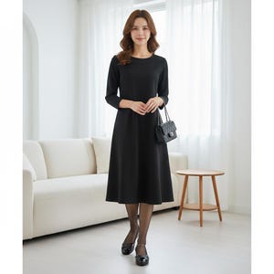 May include: A black dress with long sleeves and a round neckline. The dress has a flared skirt and is paired with black shoes and a small black handbag. The woman is standing in front of a white couch and a small wooden table.