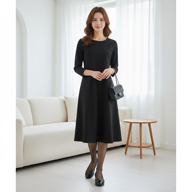 May include: A black dress with long sleeves and a round neckline. The dress has a flared skirt and is paired with black shoes and a small black handbag. The woman is standing in front of a white couch and a small wooden table.