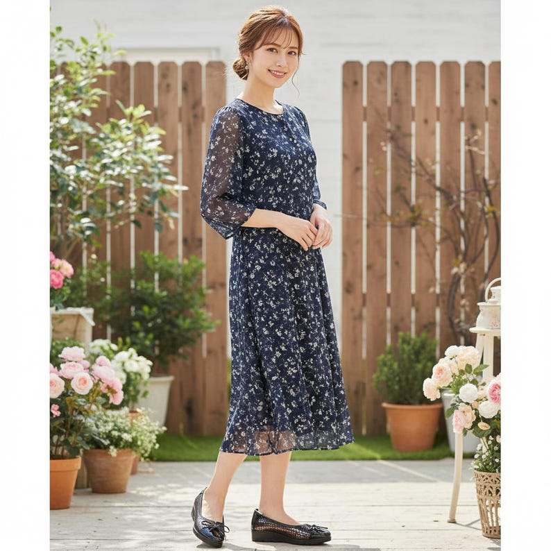 May include: A navy blue floral print dress with three-quarter sleeves. The dress features a round neckline and a flowing skirt. The model is wearing black flats. The background includes a wooden fence, potted plants, and flowers.