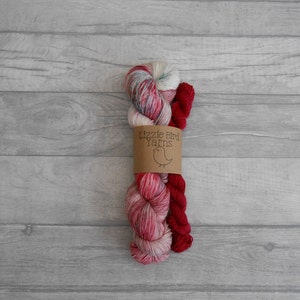 Op de afbeelding: Twee bollen garen, één rood en één met een veelkleurig patroon van roze, wit en grijs. Het garen is gewikkeld om een bruin papieren etiket met de tekst "Lizzie Bird Yarns" en een kleine rode vogel illustratie.