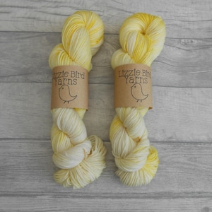 May include: Two skeins of yellow and cream hand-dyed yarn. Each skein is wrapped with a brown paper label that reads "Lizzie Bird Yarns" with a bird illustration. The yarn is displayed on a light grey wooden surface.
