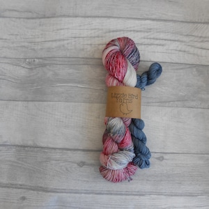 Könnte beinhalten: Ein Strang handgefärbter Wolle in verschiedenen Farben, darunter Rot, Rosa, Blau und Weiß. Die Wolle ist mit einem braunen Papieretikett mit der Aufschrift "Lizzie Bird Yarns" und einer Vogelillustration versehen. Auf einer hellgrauen Holzoberfläche.