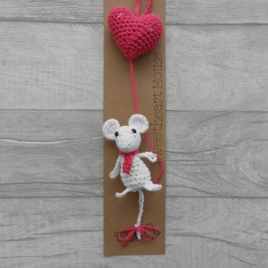 May include: A white crocheted mouse holding a pink heart balloon. The mouse is wearing a red scarf. The balloon is attached to a brown card with the text "Sweetheart Mouse".
