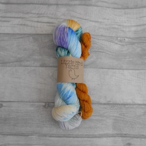 Peut inclure: Deux pelotes de laine, une bleue et blanche avec un motif moucheté et une orange unie. La laine est enroulée autour d'une étiquette en papier brun qui porte l'inscription "Lizzie Bird Yarns" avec une petite illustration d'oiseau.