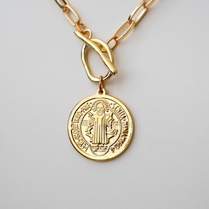 May include: A gold-colored necklace featuring a large circular pendant with an engraved religious figure. The pendant hangs from a gold chain with a toggle clasp. The necklace is displayed against a white background.